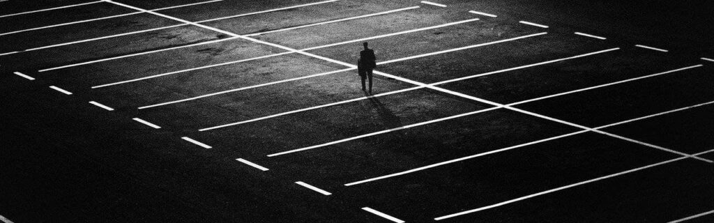 A solitary person walks in an empty parking lot at night, with bright white lines marking the spaces.